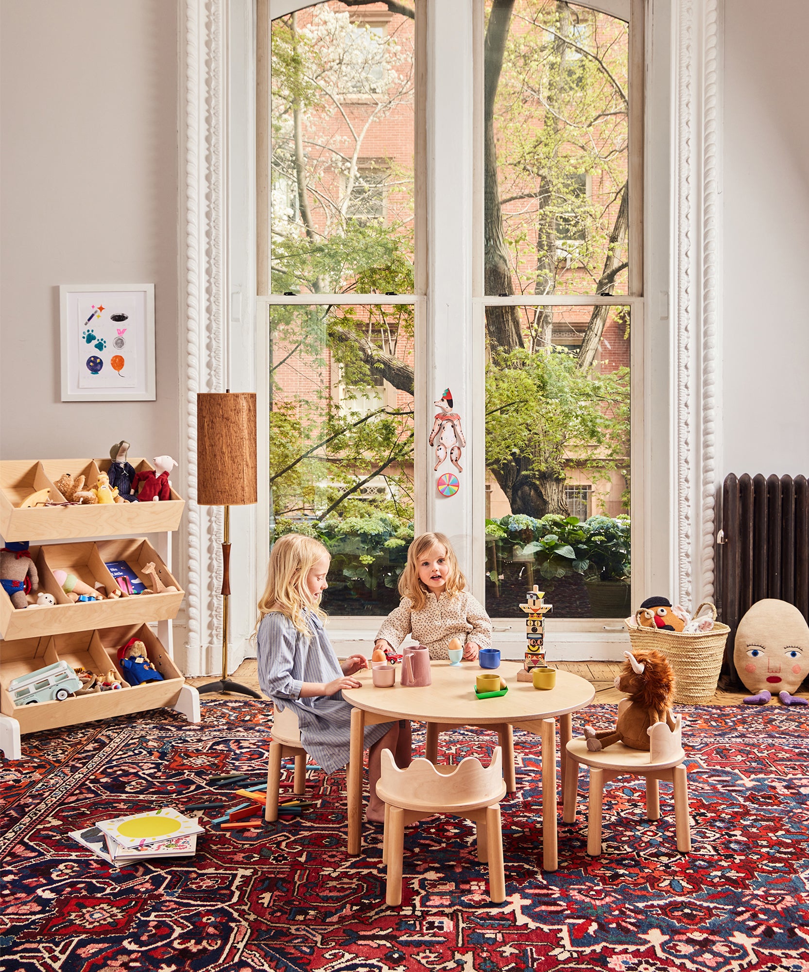 Two young children play at a small table in the Toy Store, a bright room with large windows, toys, and a patterned rug.