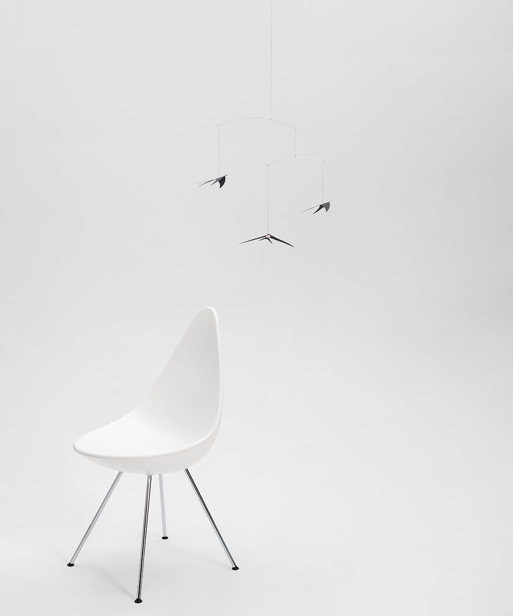 A modern white chair with metal legs sits under the Le Mobile - Flying Swallows, a black bird-shaped mobile, on a white floor against a plain white background.