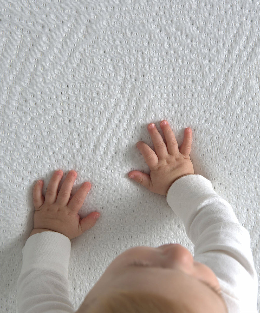 A baby in white touches the Dual Firm Crib Mattress's textured surface with both hands, viewed from above.