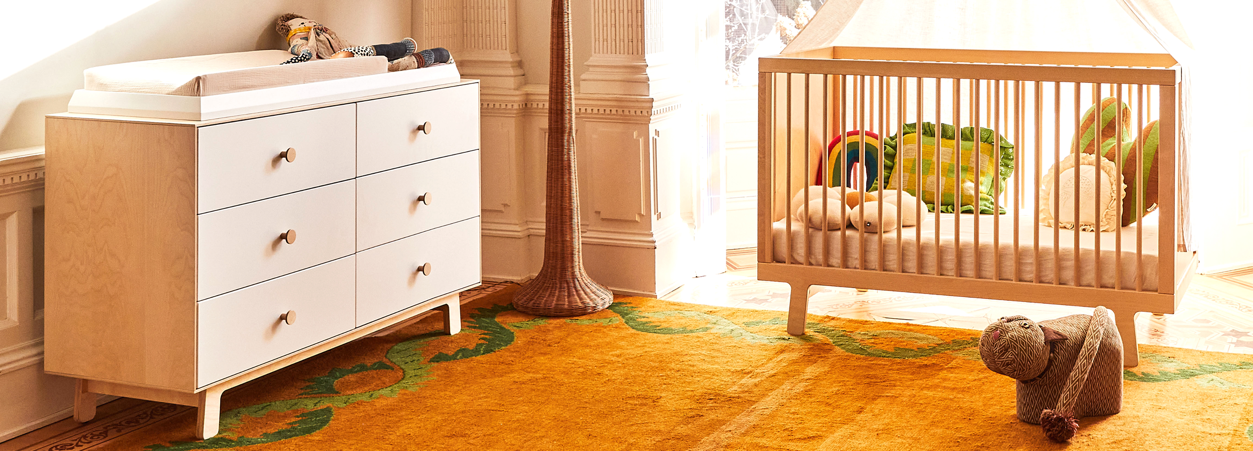 A nursery with a wooden crib, a changing table with drawers, a soft toy elephant, and a mustard yellow rug with green patterns.