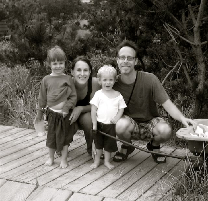 A smiling family of four poses on a wooden path surrounded by trees. Two children stand in the middle, with two adults crouching beside them. A bag is seen to the right.