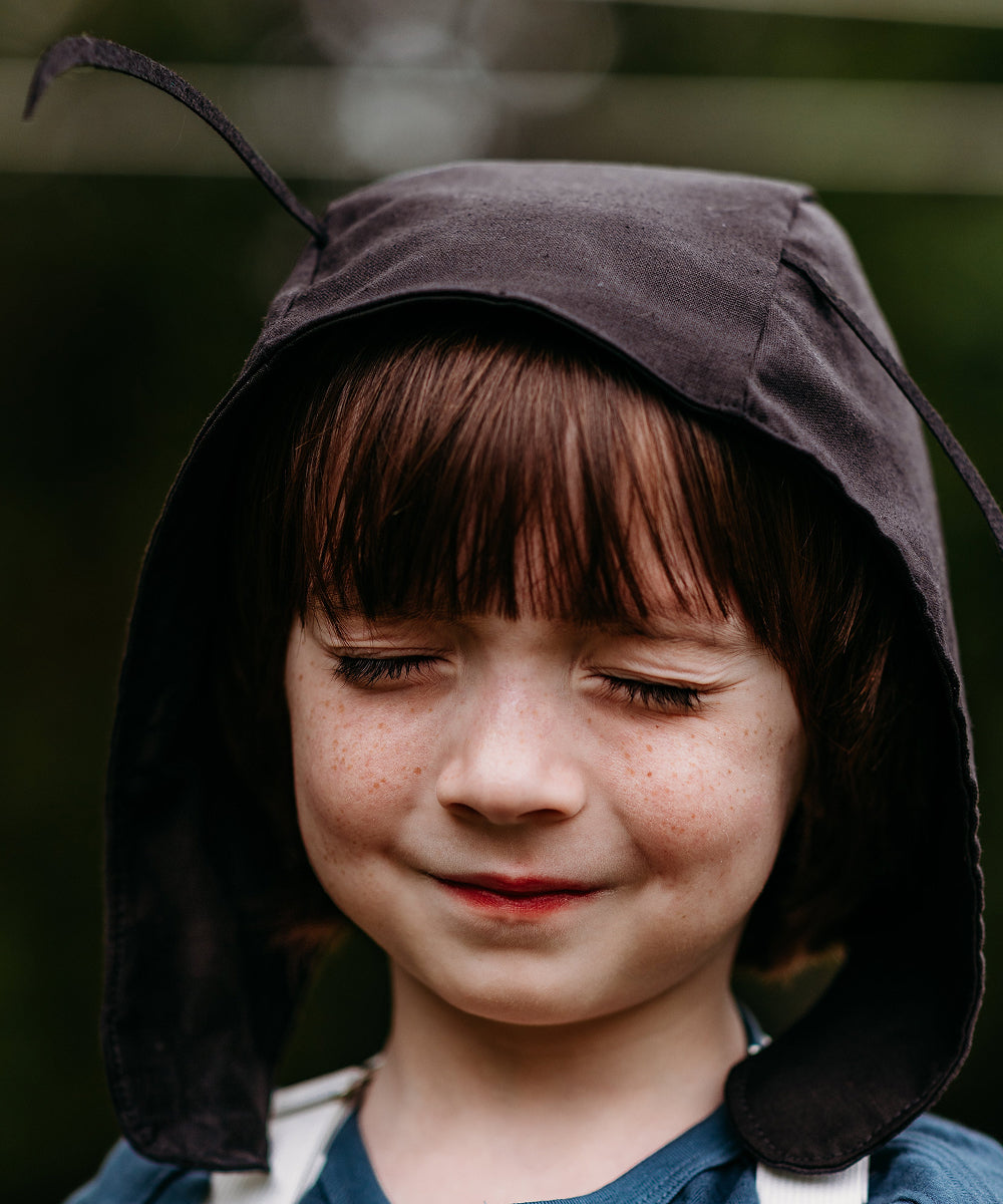 A child smiles softly with closed eyes, wearing the Le Antennae Hat—a black hood featuring ant-like antennae.
