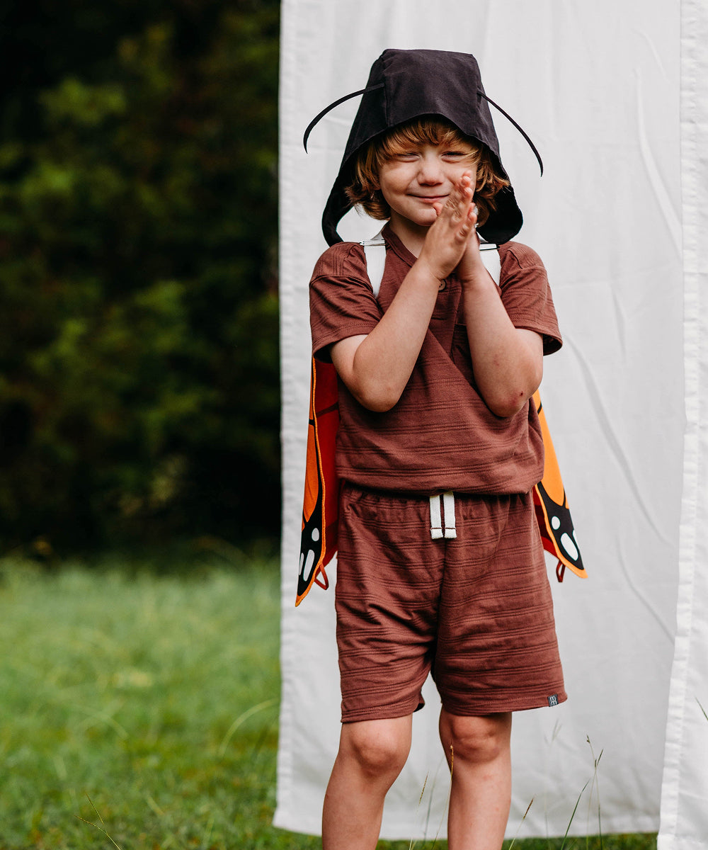 A child wearing a brown outfit, Le Antennae Hat, and butterfly wings stands smiling on grass, clapping hands; a white curtain and trees appear in the background.