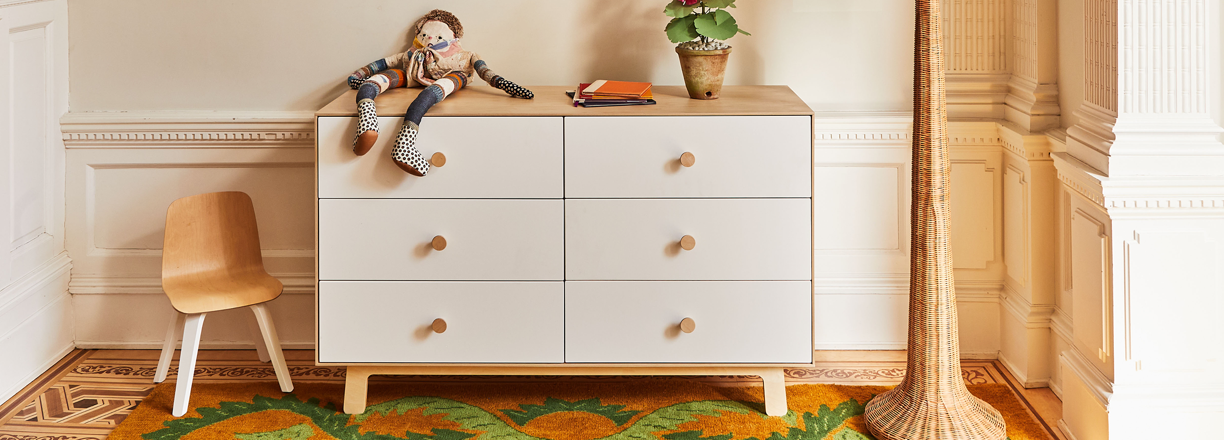 A white dresser with six drawers and round handles, a doll, and books on top. A wooden chair is on the left, and a tall woven lamp is on the right. A potted plant adds decor.