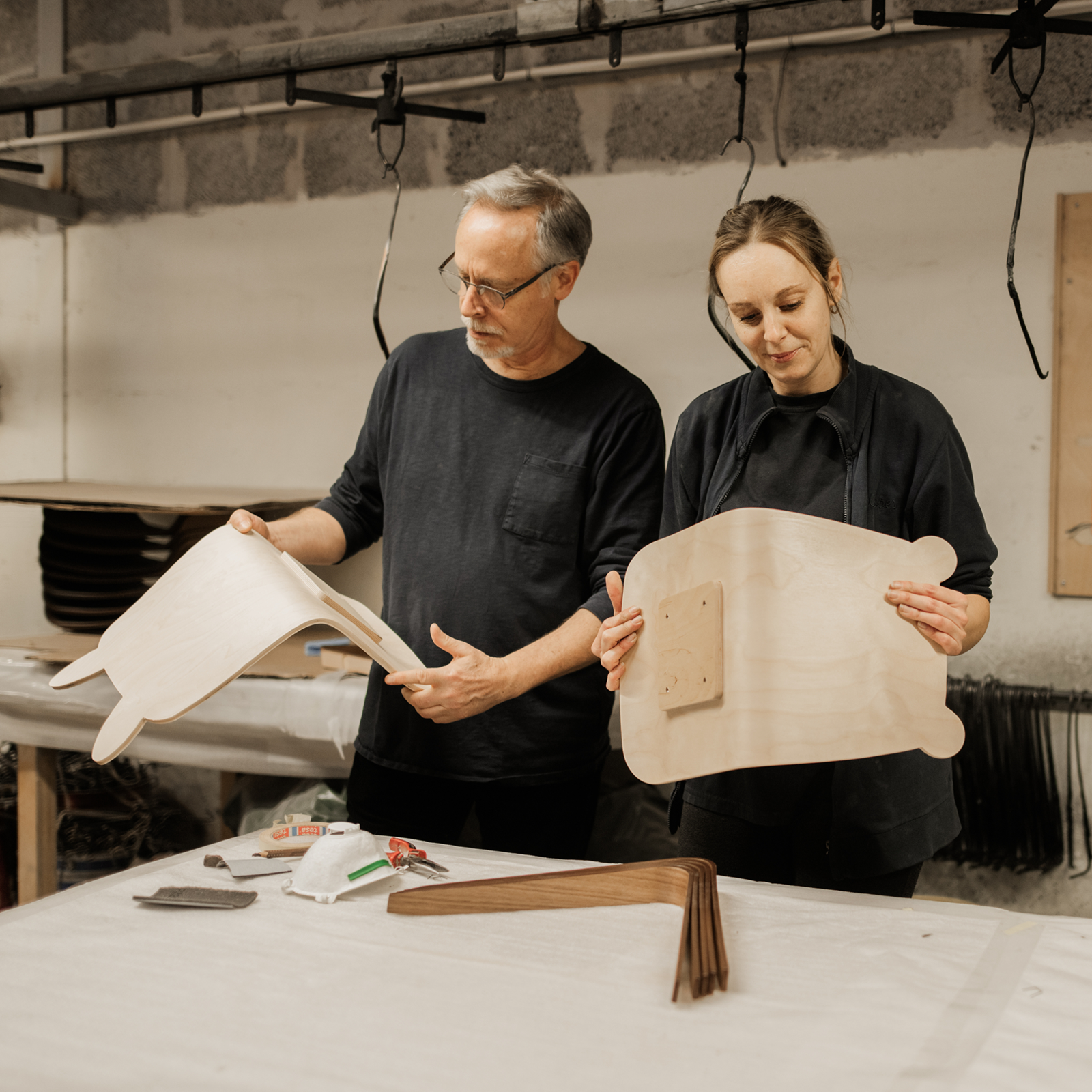 A man and a woman examine wooden chair parts in a workshop.