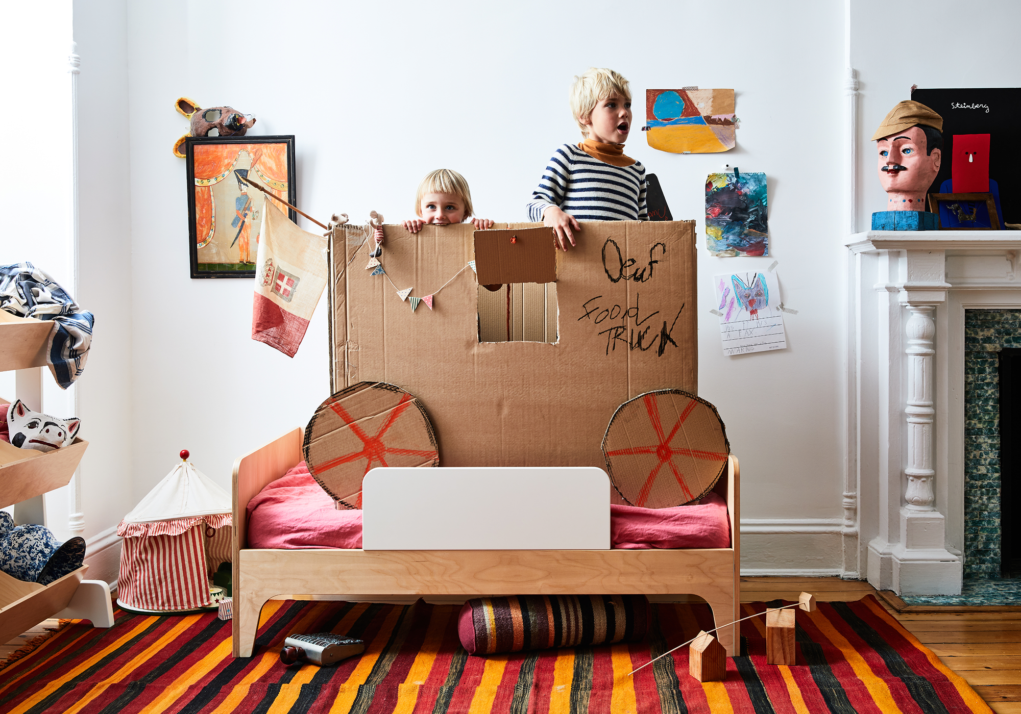 Children playing in a room with a cardboard car on a bed. Artwork and toys are visible. A striped rug covers the wooden floor.