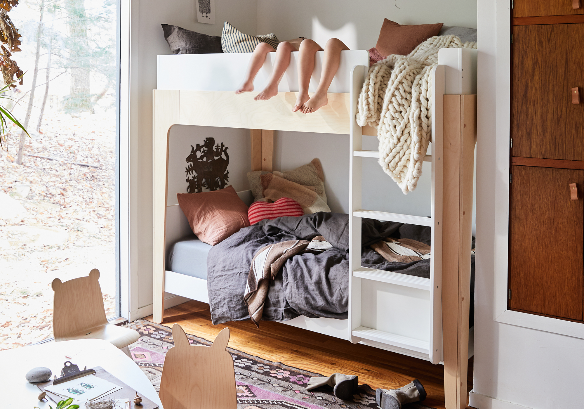 A cozy bedroom with a white bunk bed. Two children’s feet hang from the top bunk. Blankets and pillows are on both beds, and a rug with a play table is on the wooden floor.