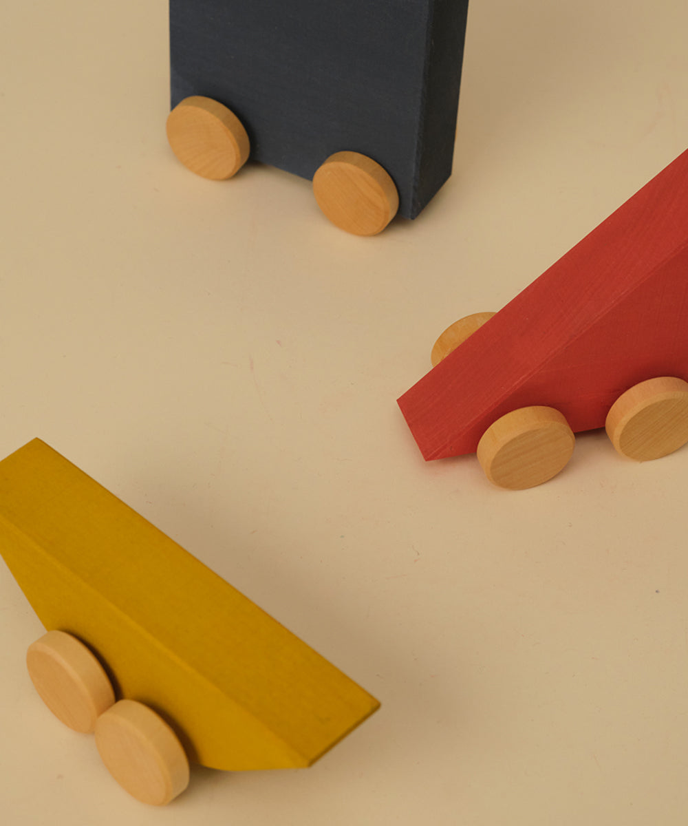 The Le Vehicle Trio features three colorful wooden toy cars—yellow, red, and blue—displayed on a beige surface from above.