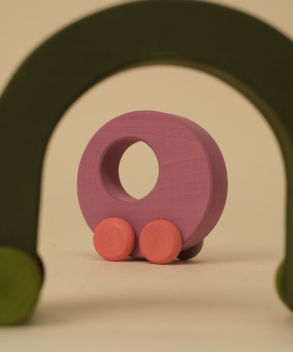 Le Vehicle Duet is a purple wooden toy with a hollow center, pink wheels, and a green arch in the foreground.