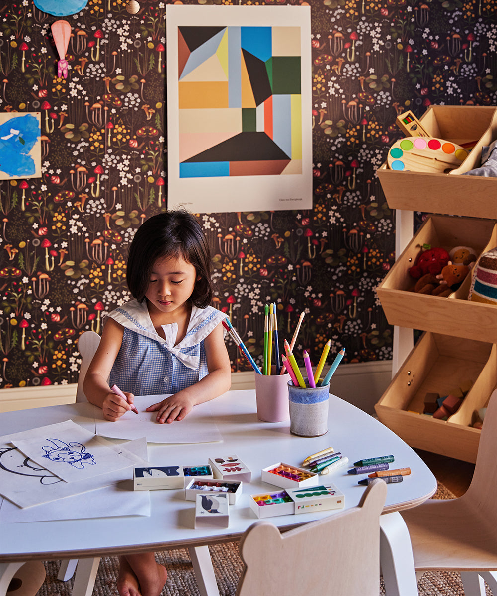 A young girl sits at a table drawing with markers, surrounded by crayons and papers. Colorful, patterned wallpaper adorns the room, along with art supplies on shelves and a Le Collage One Poster adding charm to the setting.