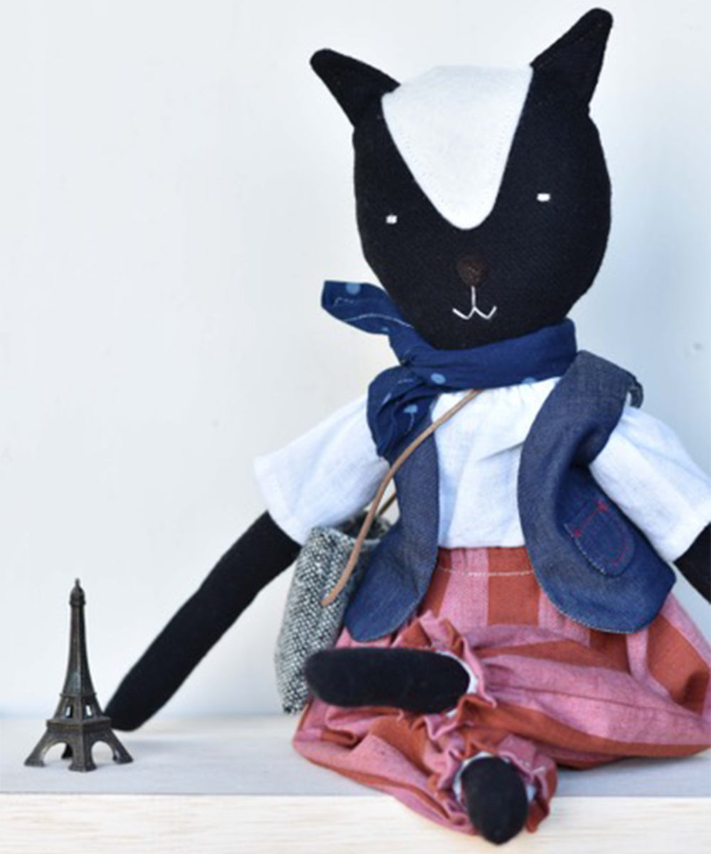 Theo le Skunk, wearing a skirt and scarf, sits beside a small Eiffel Tower figurine on a light surface.