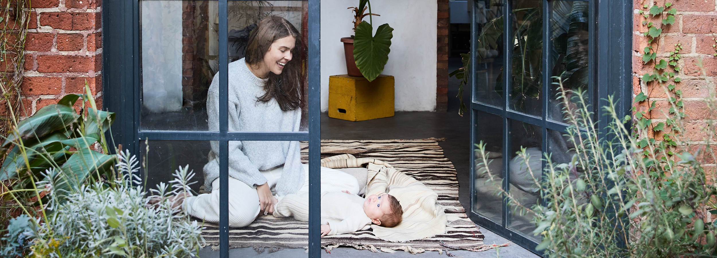 Woman sitting on a rug smiles at a baby lying beside her. They are in a room with glass doors and surrounded by plants.