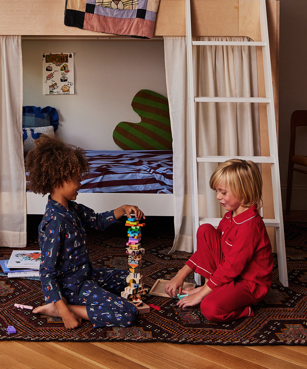 Two children in pajamas play with the Le Totem Stacker toy on the floor of a bedroom with a bunk bed.