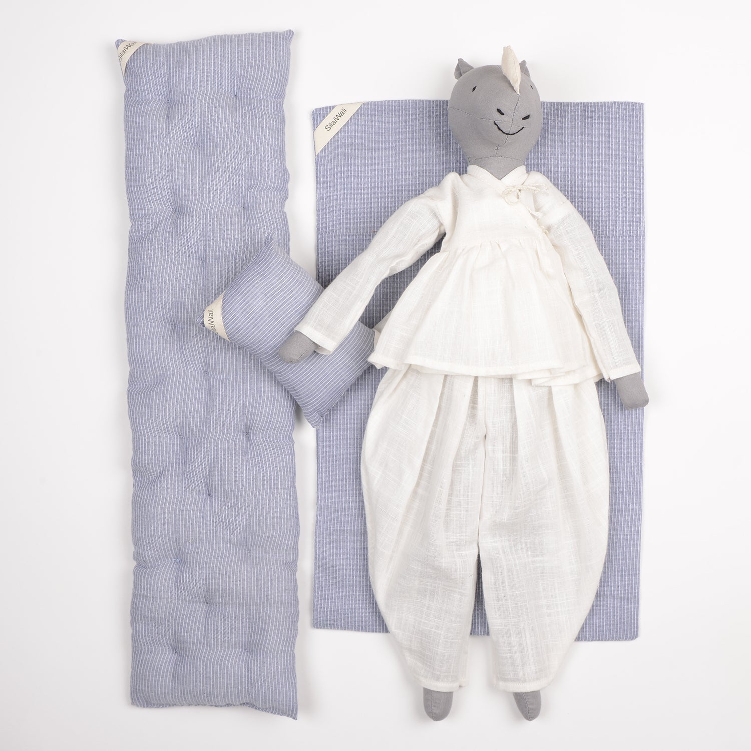 Le Rhino, a plush toy with a unicorn horn, is dressed in white pajamas and rests on a blue mattress with a matching pillow against a white background.