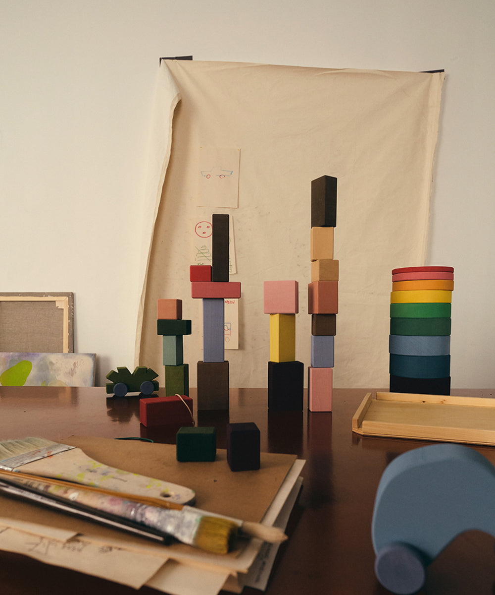 Le Rectangles, vibrant wooden stacking blocks, are displayed on a table with art supplies and a canvas backdrop.