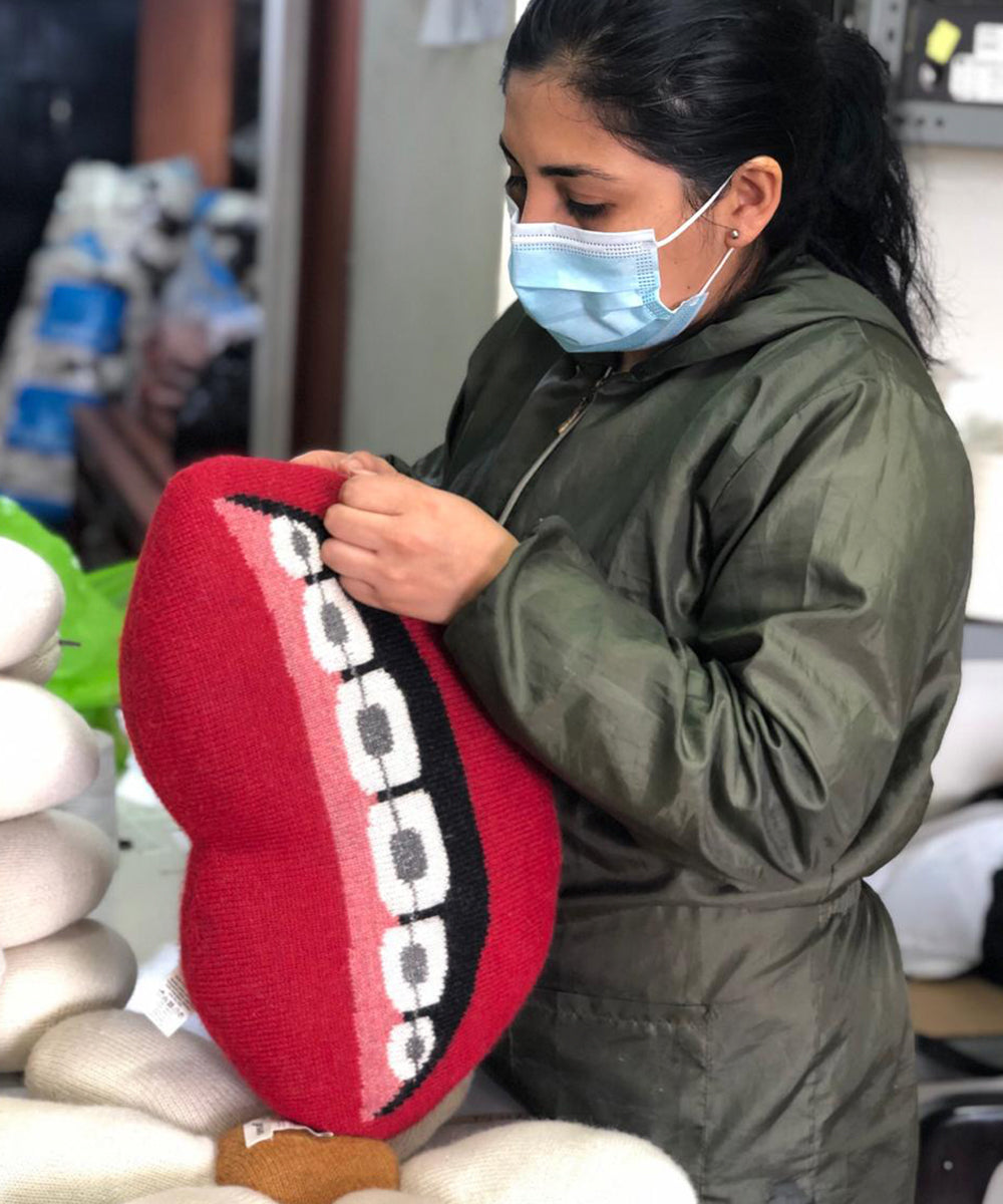 A masked individual sews a Mouth Pillow - Braces, designed as a red pillow shaped like a mouth with teeth.