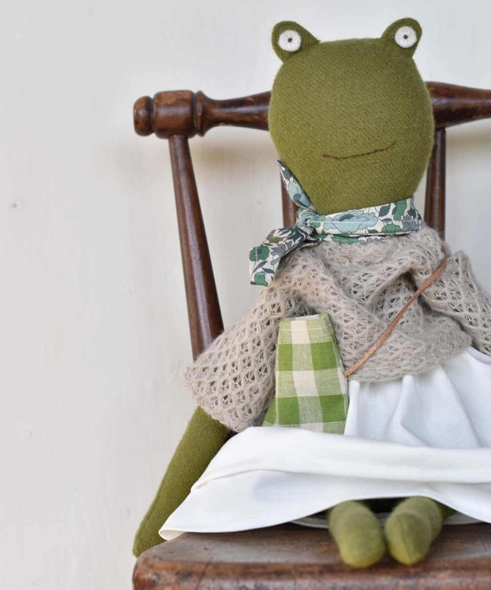 Fern le Frog, a plush frog doll dressed in a scarf, dress, and shawl, sits on a wooden chair against a plain wall.