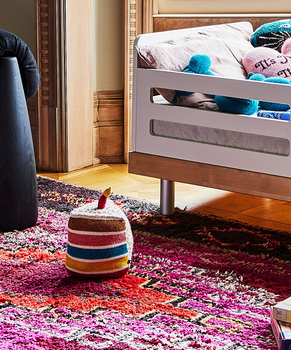 A colorful children's room features a bed with a Birthday Cake Pillow, a striped pouf, and a vibrant rug, complete with stuffed animals on the bed.