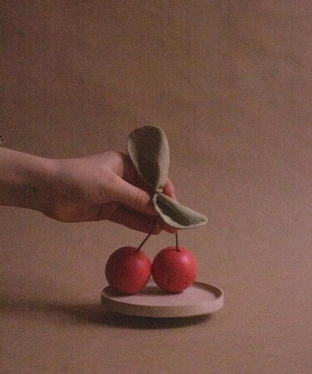 A hand holds the Le Wooden Cherry Play Set, featuring artificial cherries with cloth leaves, above a small round dish on a brown surface.