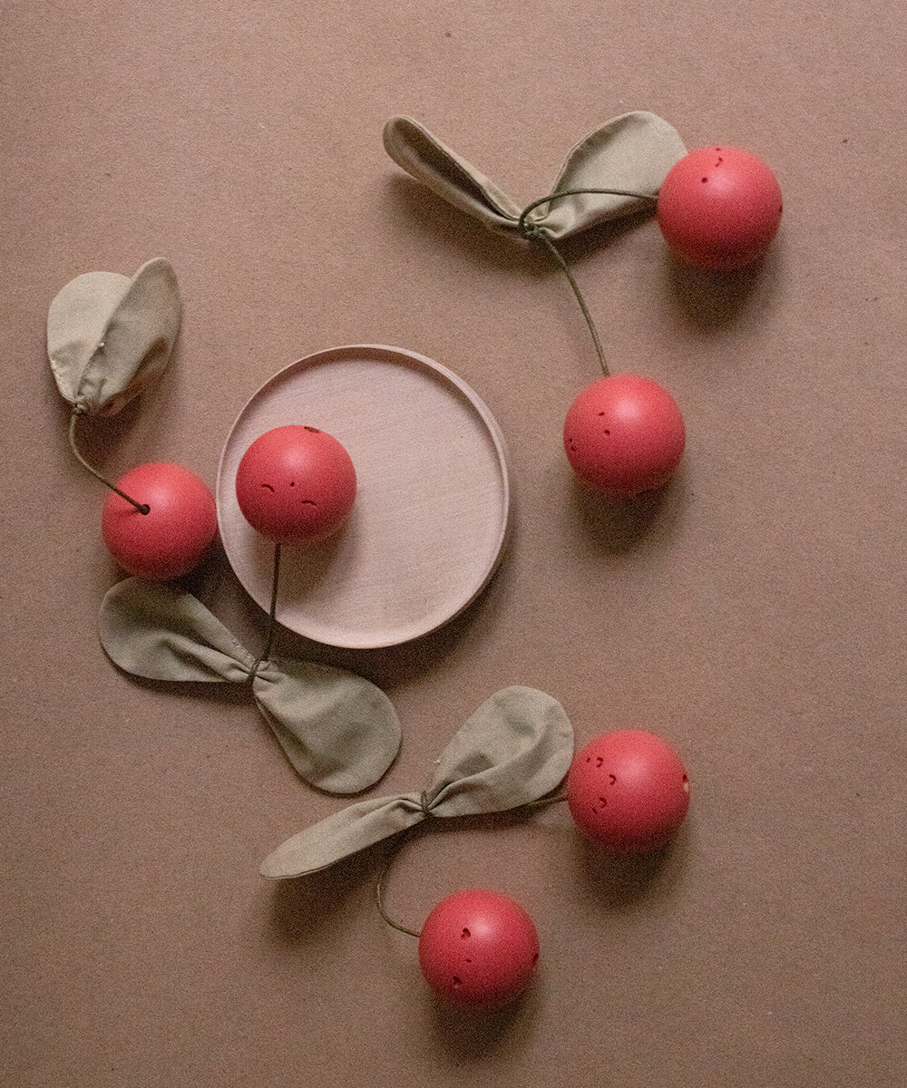 The Le Wooden Cherry Play Set features a tan surface adorned with five red artificial cherries connected to green fabric leaves, encircling a round, empty light brown dish in the center.