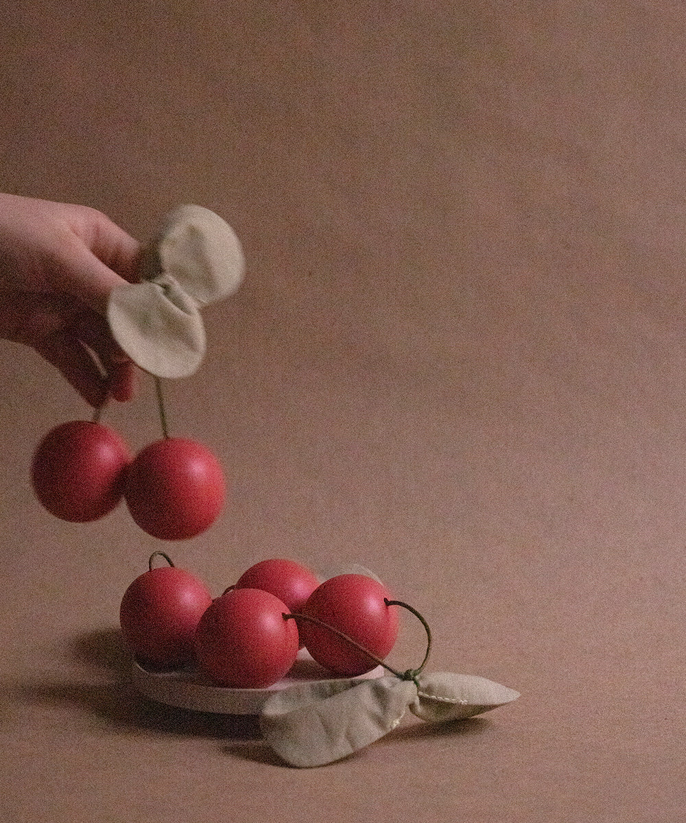 A hand holds a pair of cherries and leaves from the Le Wooden Cherry Play Set, above a dish with more play cherries.