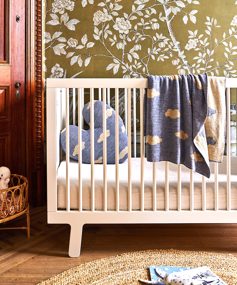 A floral wallpaper backdrop showcases a white crib adorned with cloud-inspired pillows and the Cloud Blanket, while a woven basket sits on the floor.