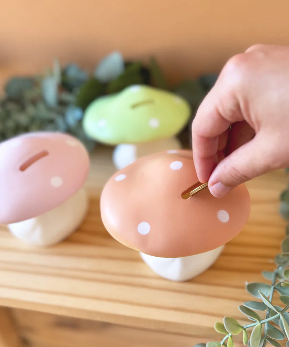 A hand places a coin into the Le Mushroom Bank, a mushroom-shaped piggy bank, on a wooden surface with plants in the background.