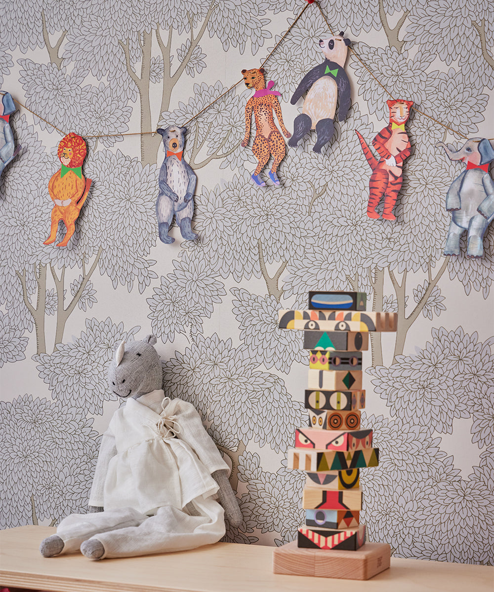 A plush toy dressed in white sits on a wooden surface next to vibrant stacked blocks, with Le Garland - Animal Parade cutouts dangling from strings against tree-patterned wallpaper.