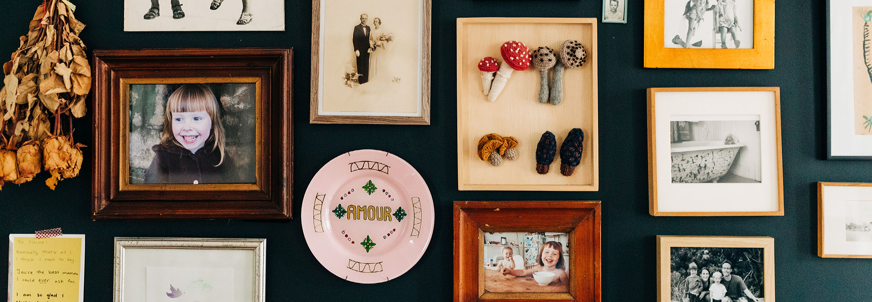 A wall collage featuring framed family photos, a decorative plate with AMOUR, dried flowers, and fabric mushrooms in a box.