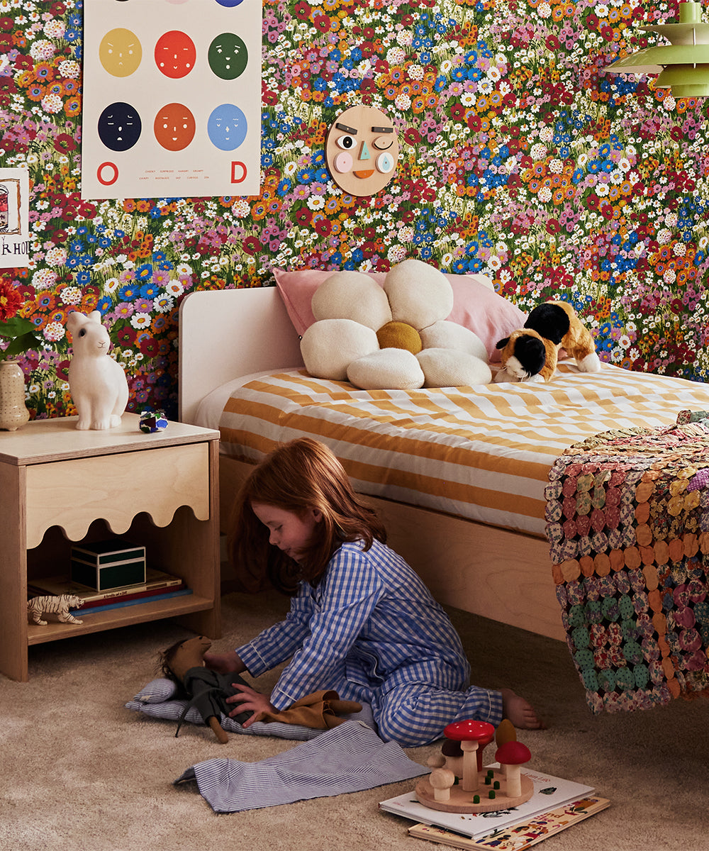 A child in pajamas plays with the Le Mushroom Sorter on a carpeted floor in a colorful floral-themed bedroom, featuring a wooden bed and various toys.