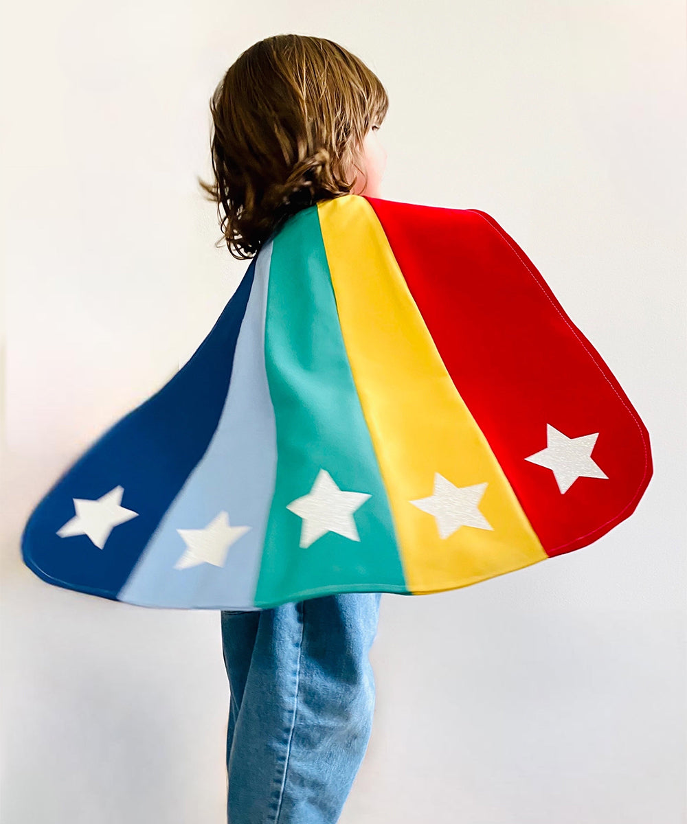 A child in the Le Superhero Cape adorned with stars stands sideways against a plain background.