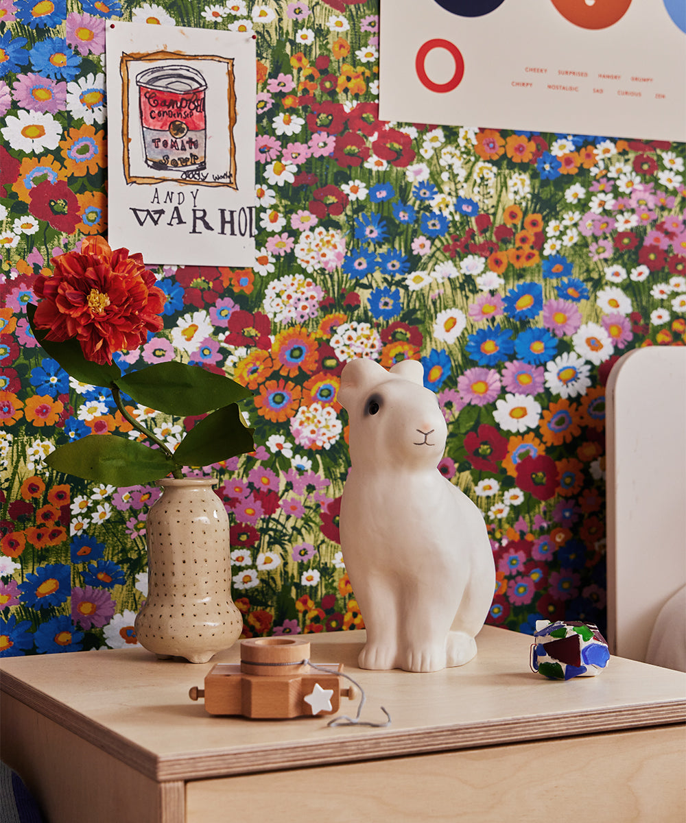 A colorful floral wallpaper serves as a backdrop for the Le Bunny Lamp, featuring a white rabbit figurine, alongside a flower in a vase, a toy, and some posters on a wooden table.