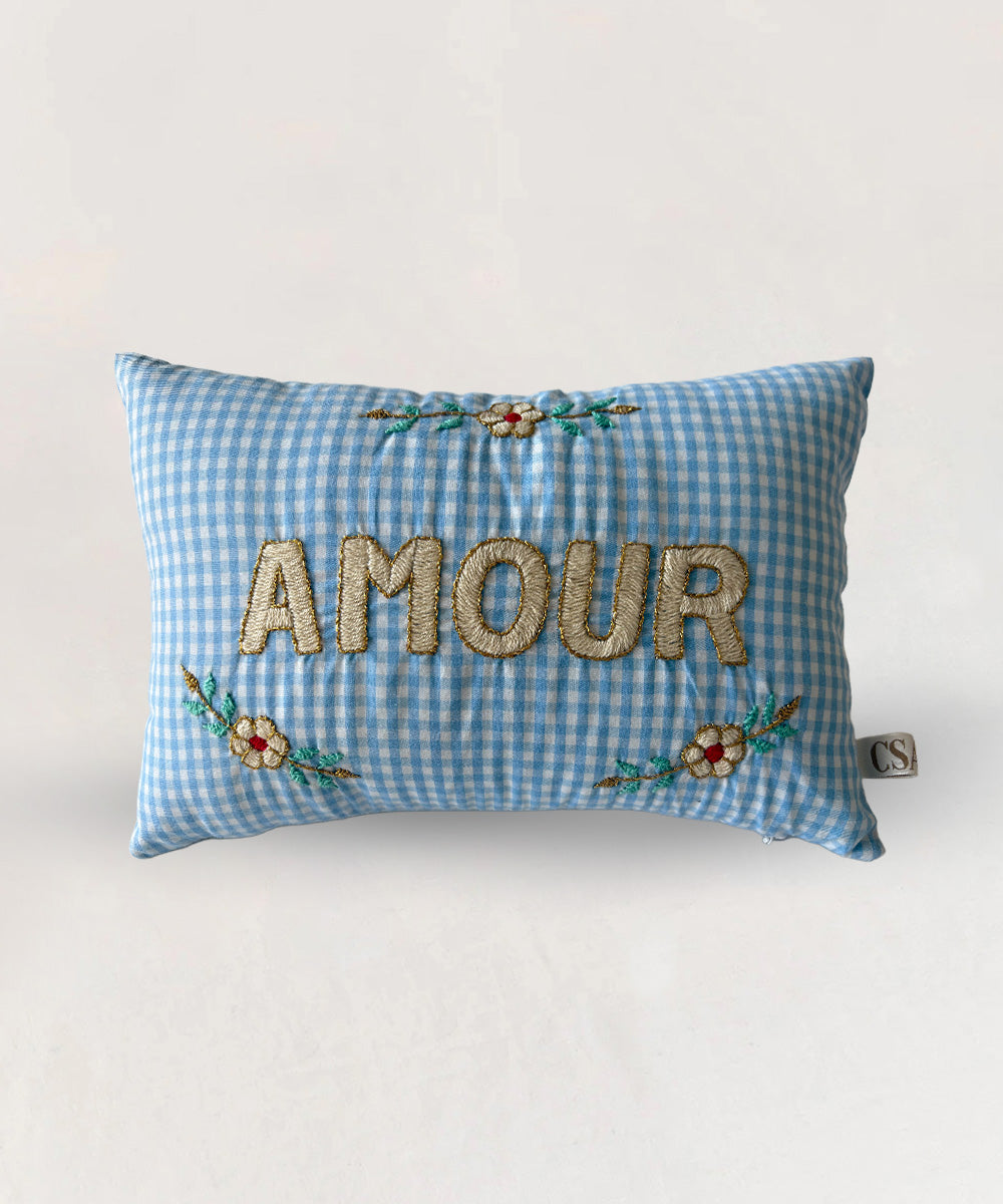 Le Mini Embroidered Pillow in blue gingham features "AMOUR" and floral embroidery in gold, red, and green threads.