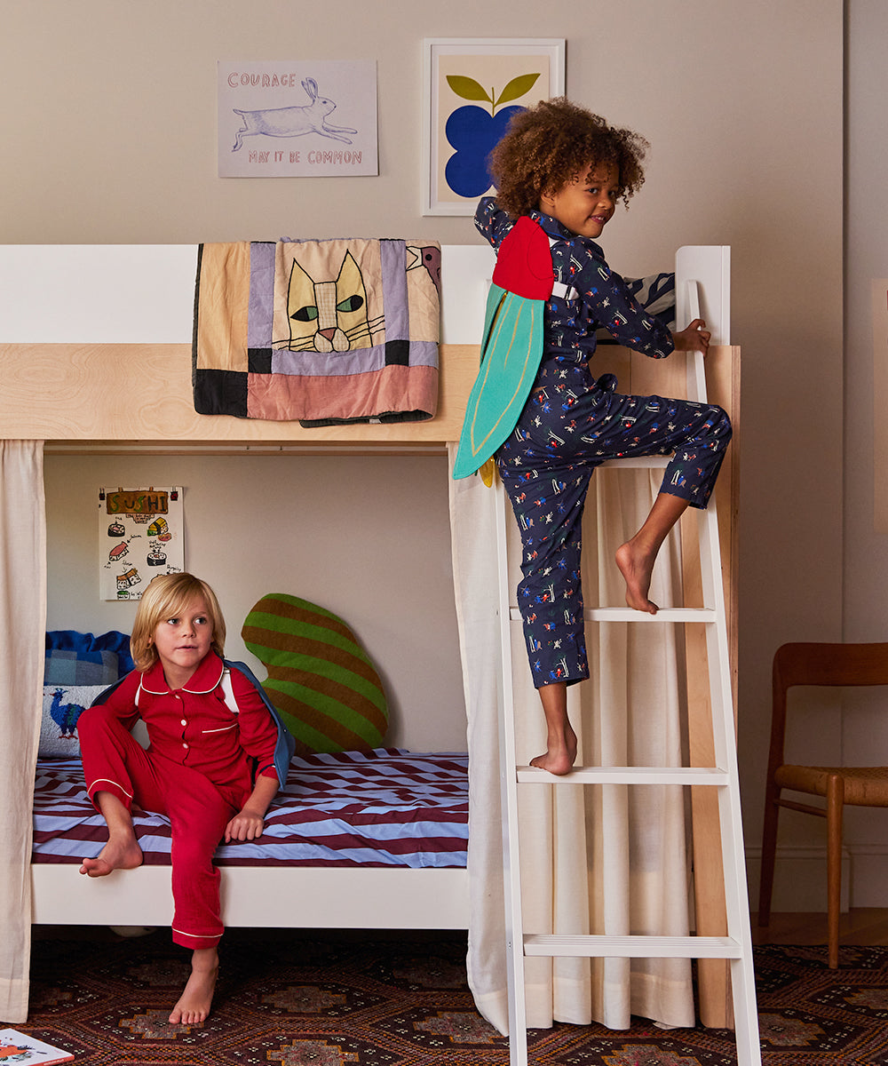 Two children in pajamas play around a bunk bed; one climbs a ladder with a Le Firefly Costume attached to their back, while the other sits on the bottom bed. The room is decorated with paintings and quilts.