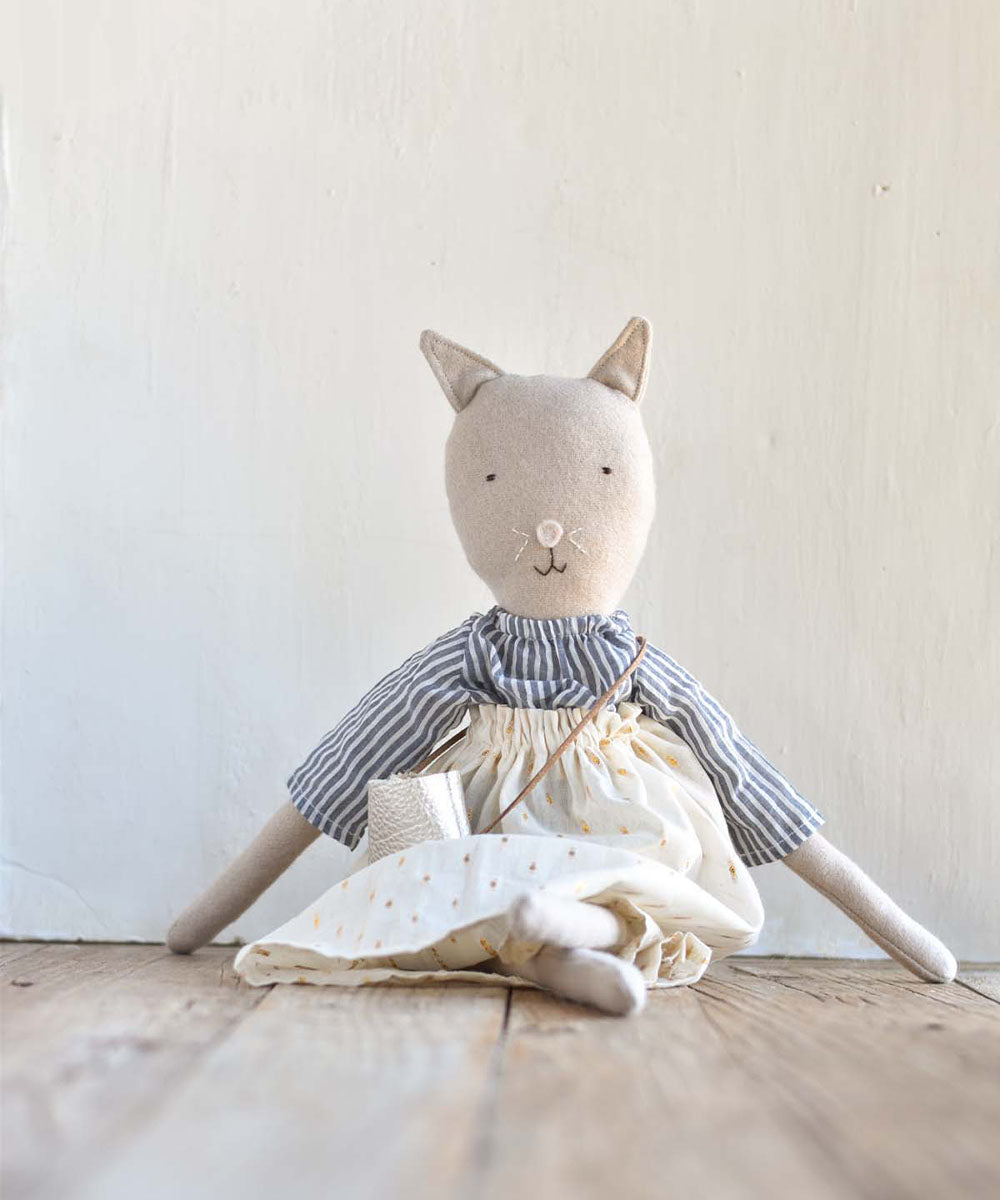 Mae le Cat – Field Skirt: Stuffed cat doll in a striped shirt and cream skirt sitting on a wooden floor against a light wall.