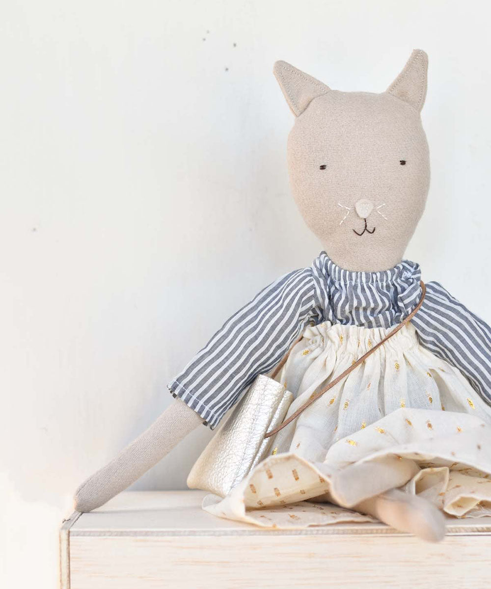 Mae le Cat – Field Skirt: Stuffed cat doll in a striped shirt, white skirt, and purse, seated on a wooden surface.