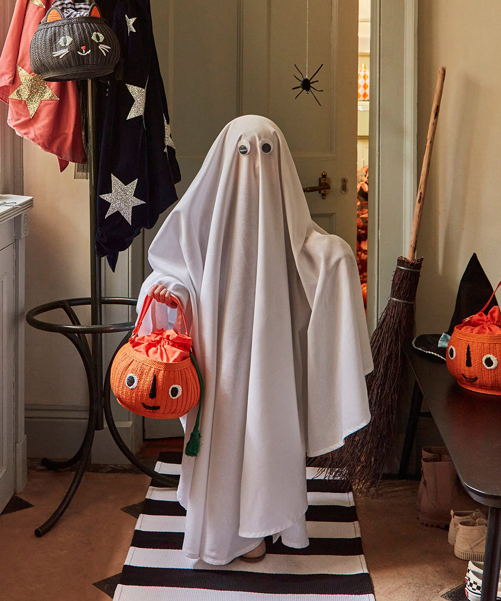 A child in a ghost costume with googly eyes holds the Le Pumpkin Basket Bag while standing in a decorated hallway.