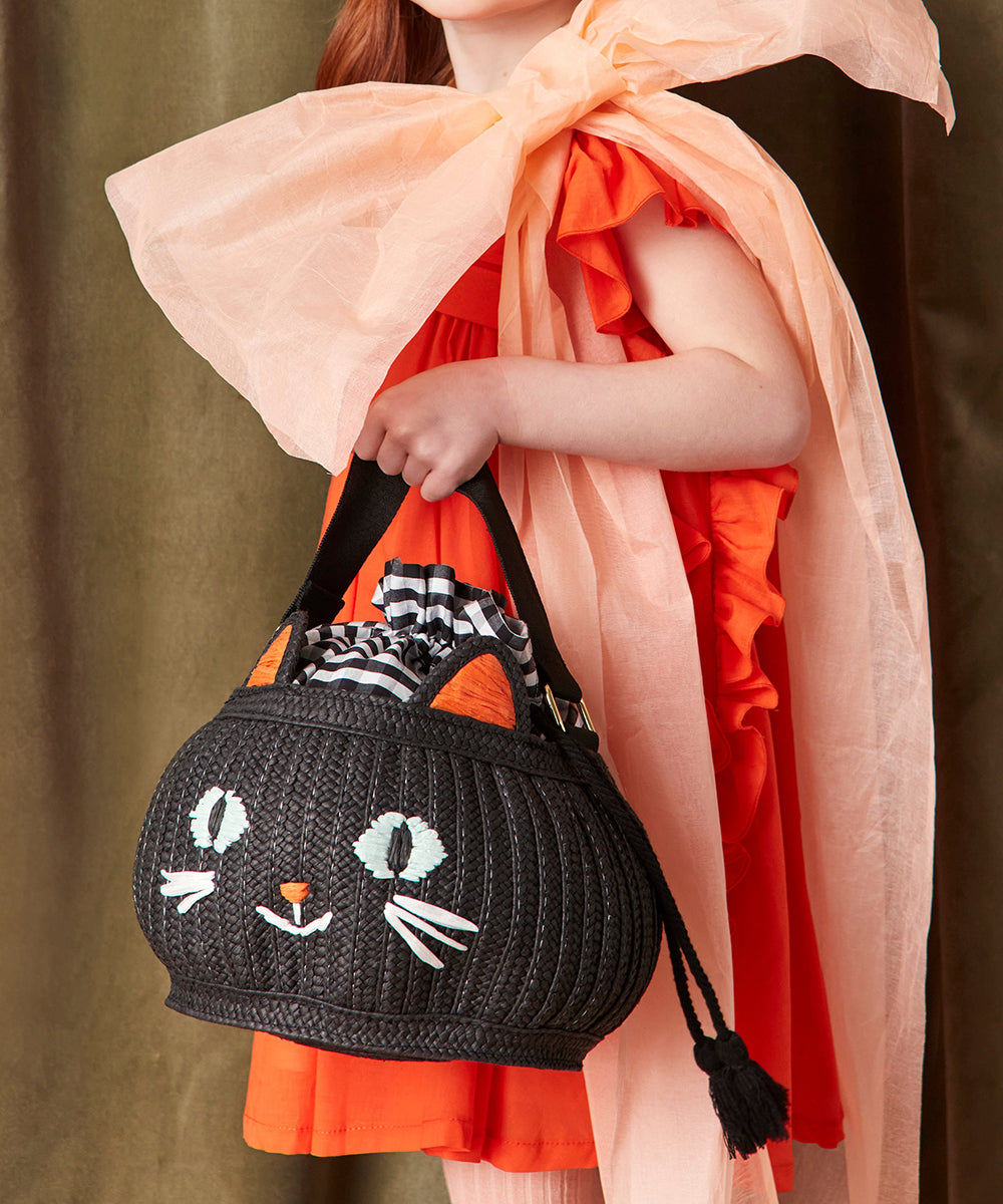 Wearing an orange dress, the girl holds the Le Cat Basket Bag—a black cat-shaped bag with orange ears, white face details, and a peach bow draped over her shoulders.