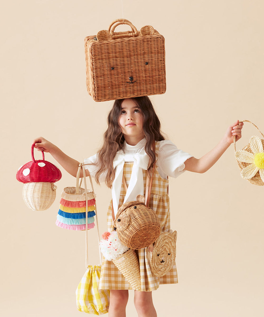 Girl in a yellow gingham dress holds woven bags, with a Mushroom Basket balanced on her head.