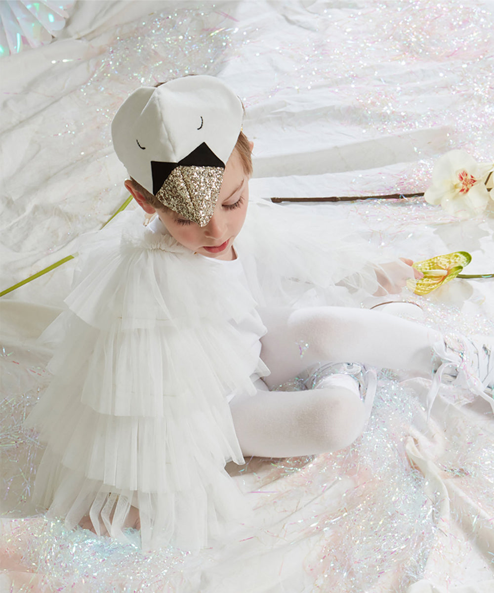 A child wears the Swan Costume, featuring a ruffled cape and beak hat, while sitting on a shimmery white blanket.