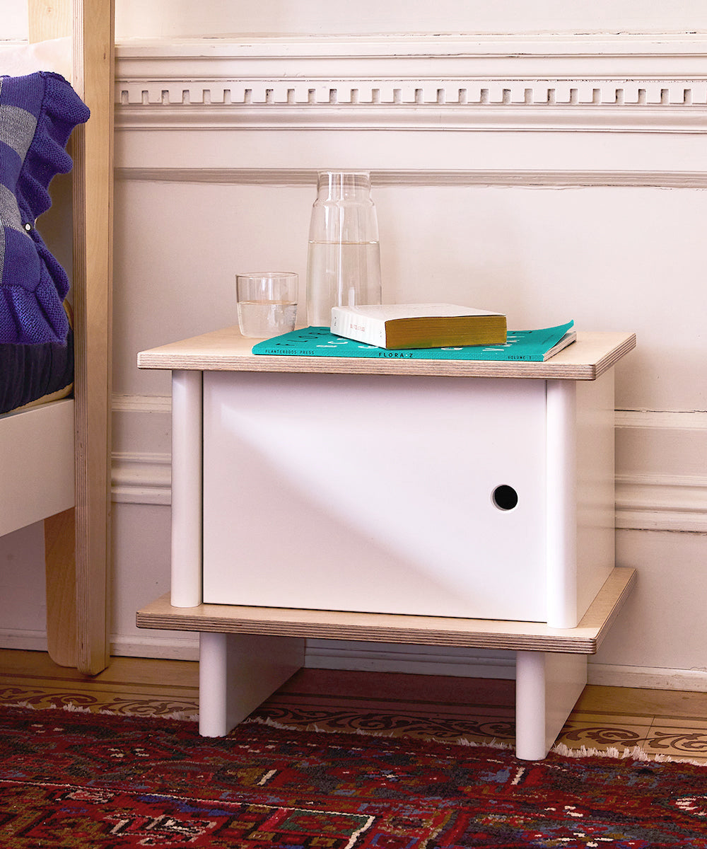 The ML Nightstand is shown with a glass, carafe, book, and magazine on top, placed beside a bed and a patterned rug.