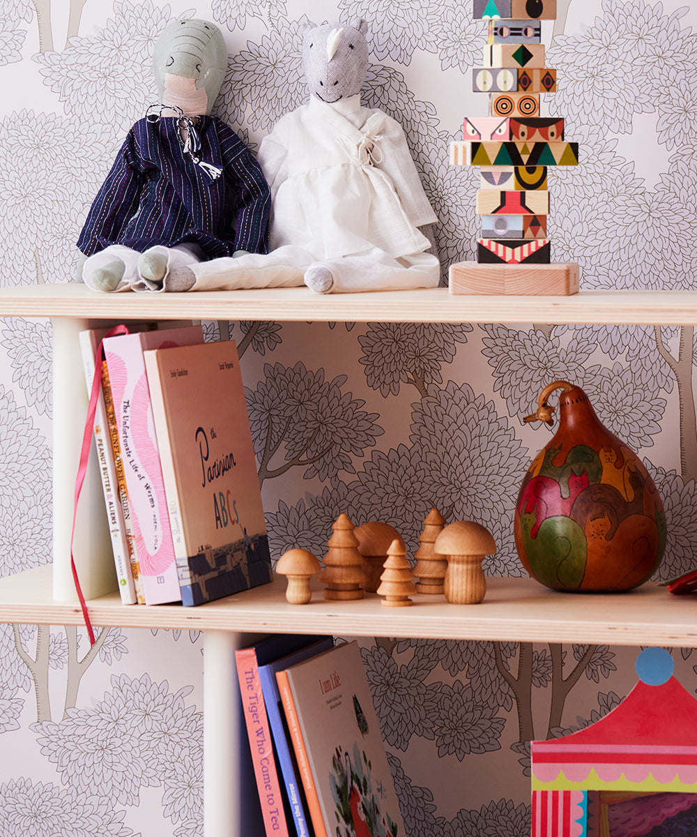 Children's room shelf with plush toys, colorful books, Le Wooden Mushroom & Tree Set, and a painted gourd against floral wallpaper.