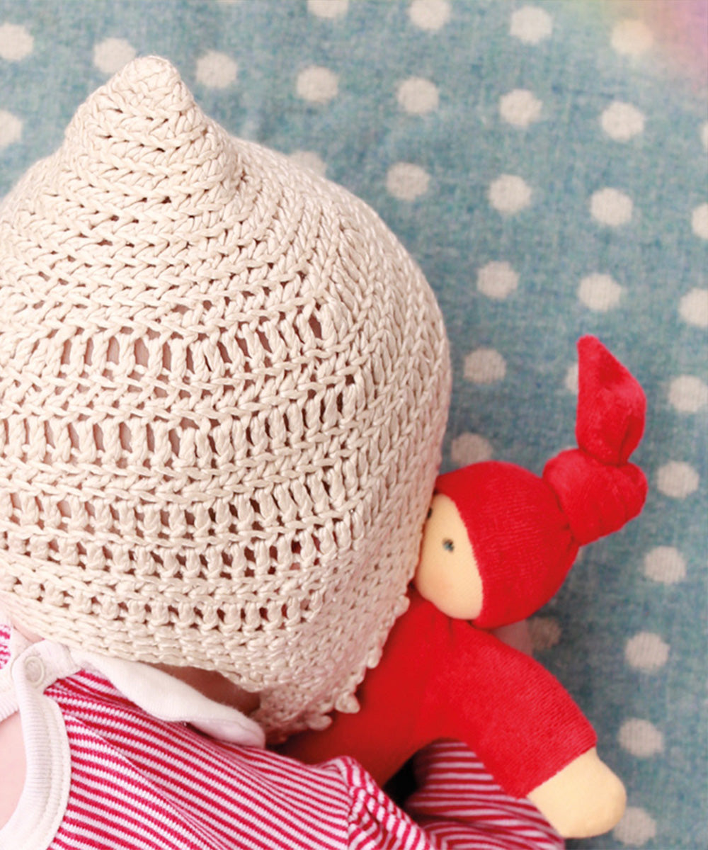 Baby cuddling a Waldorf Doll - Red, wearing a knit hat and lying on a blue blanket with white polka dots.