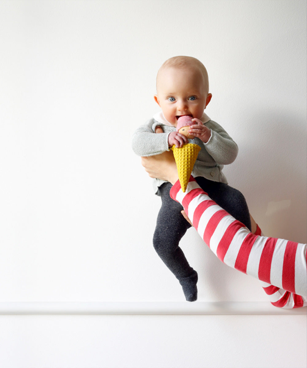 A baby licks an Ice Cream Rattle toy while being held by an adult in a red and white striped sleeve.