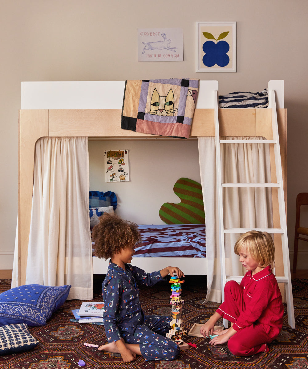 Two children in pajamas play with blocks in a bedroom featuring a Perch Twin Bunk Bed, curtains, and colorful wall decorations.