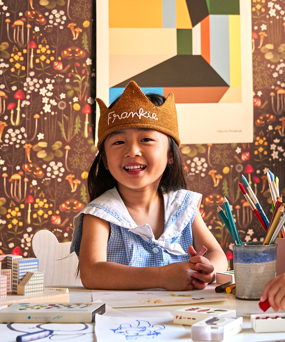 A smiling child wearing a white Personalized Gold Crown labeled "Frankie" sits at a desk drawing in a colorful, decorated room.