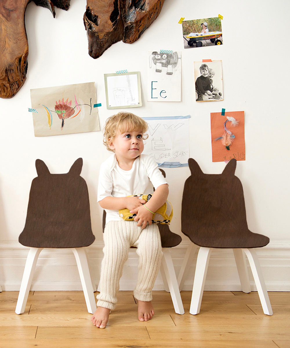 A toddler sits on a low chair between two dark-brown wooden Bunny Play Chair (Set of 2) chairs with rabbit-ear backrests.
