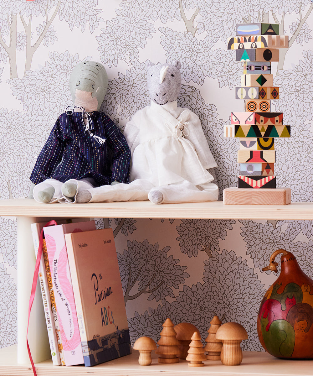 Shelf featuring two fabric dolls, a Le Rhino wooden stacking toy, small wooden cones, books, and a decorative gourd set against floral wallpaper.