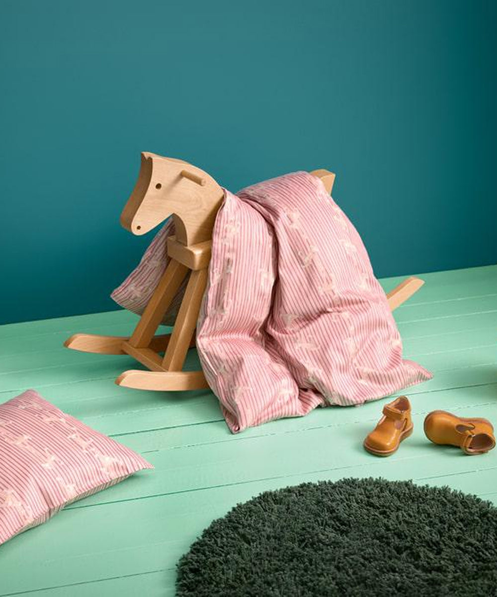 The Kay Bojesen Rocking Horse with a pink blanket stands beside a matching pillow, brown shoes, and a green rug on a mint-green floor and teal wall backdrop.