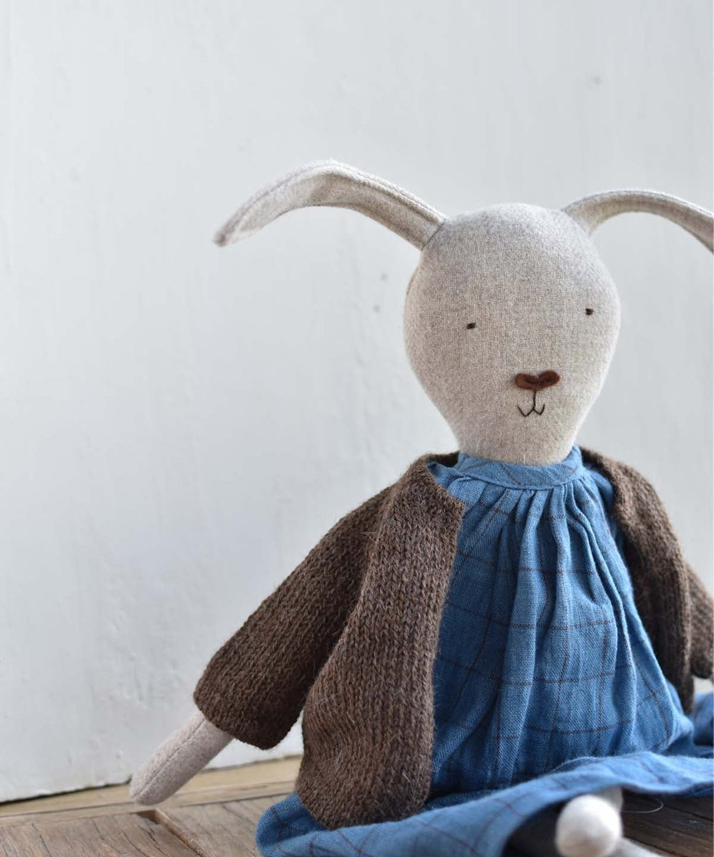 Ruthie le Rabbit, a stuffed toy with long ears in a blue dress and brown knit sweater, sits on a wooden floor by a white wall.